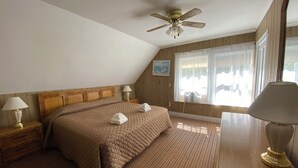 Un dormitorio con cabecera de madera, cama con colcha marrón, dos lámparas de noche, ventilador de techo, ventana con cortinas traslúcidas y un cuadro pequeño en la pared.