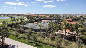 Un complexe de courts de tennis comprenant plusieurs terrains, entouré de palmiers et offrant une vue sur un lac et des collines lointaines.