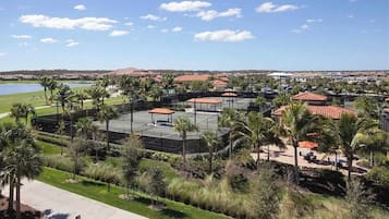 A tennis court complex with multiple courts, surrounded by palm trees and a view of a lake and distant hills.
