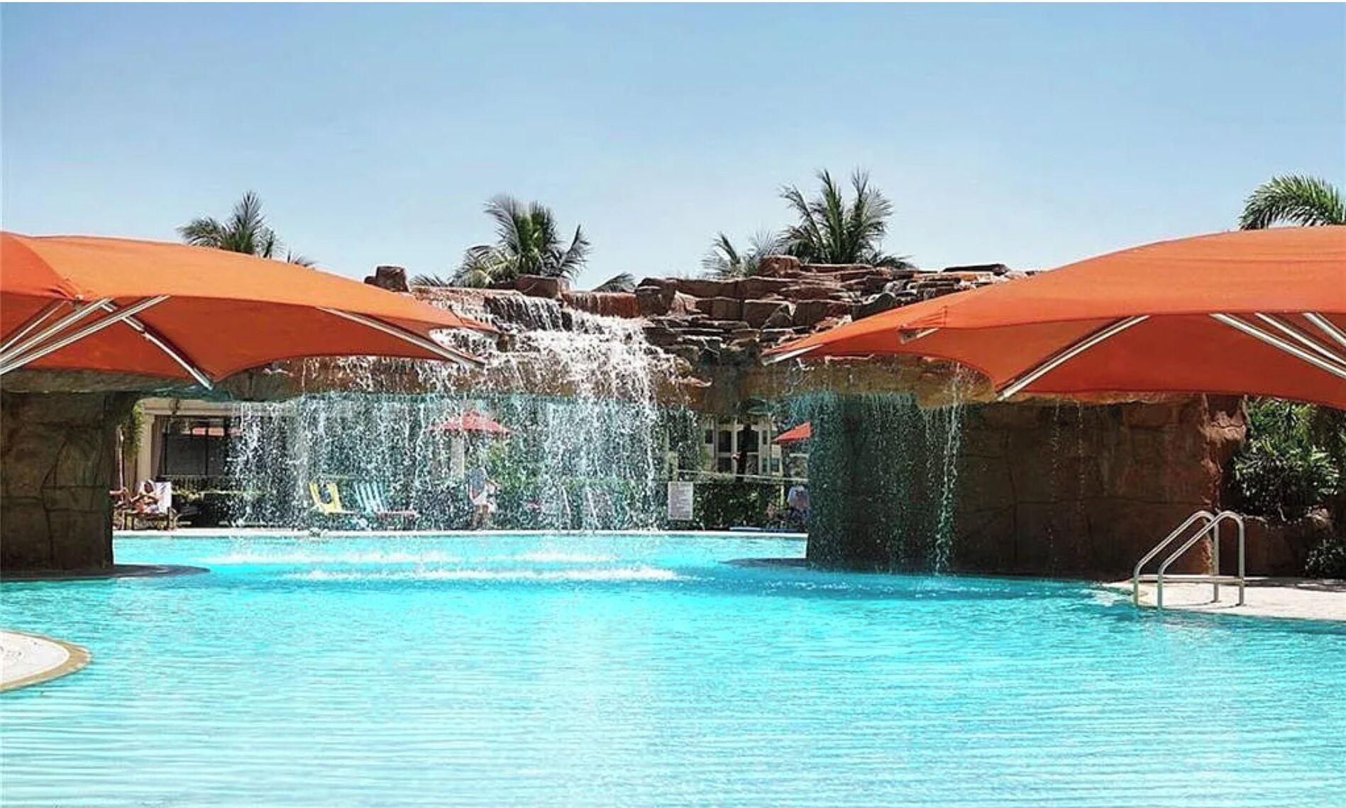A swimming pool with a waterfall feature, surrounded by palm trees and covered by orange umbrellas.