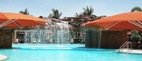 En swimmingpool med ett vattenfall, omgiven av palmer och med orangefärgade parasoller som ger skugga.