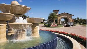 Une fontaine à plusieurs niveaux, avec de l’eau jaillissant du niveau supérieur. En arrière-plan, on aperçoit un bâtiment doté d’une entrée en arc de cercle et un parking avec des voitures.
