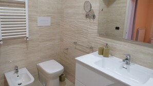 Et badeværelse med toilet, bidet, vask, spejl og håndklæderække.
