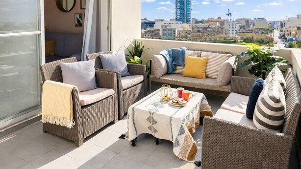 Un balcon agrémenté de meubles en osier, une table recouverte d’une nappe et une vue sur le paysage urbain.