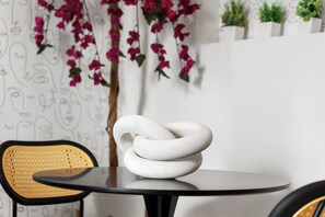 Uma mesa moderna com uma escultura branca em espiral como peça central, uma cadeira de vime e uma parede com um padrão de rostos e plantas.