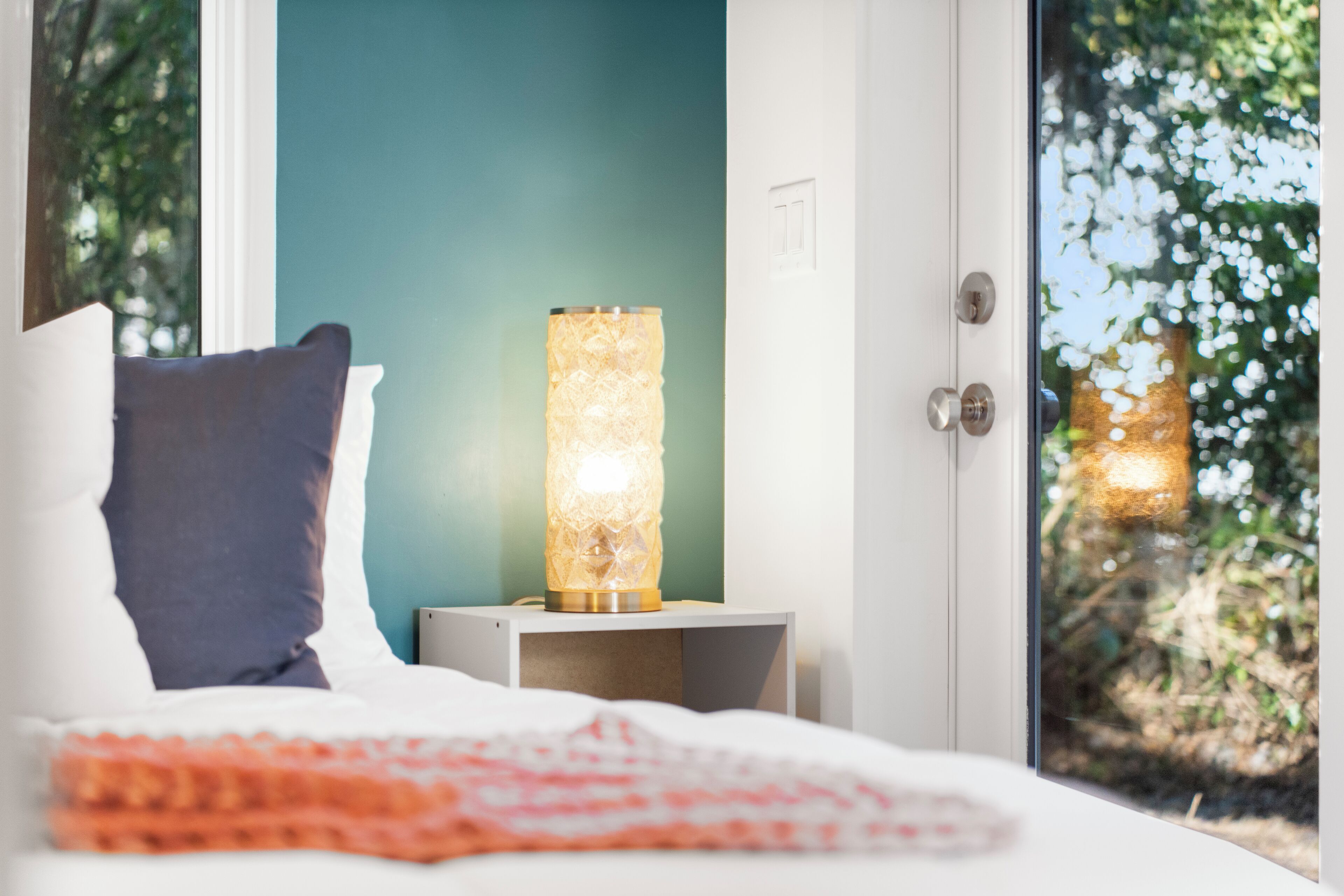 A bedroom with a bed, a nightstand with a lamp, and a window with a view of trees.