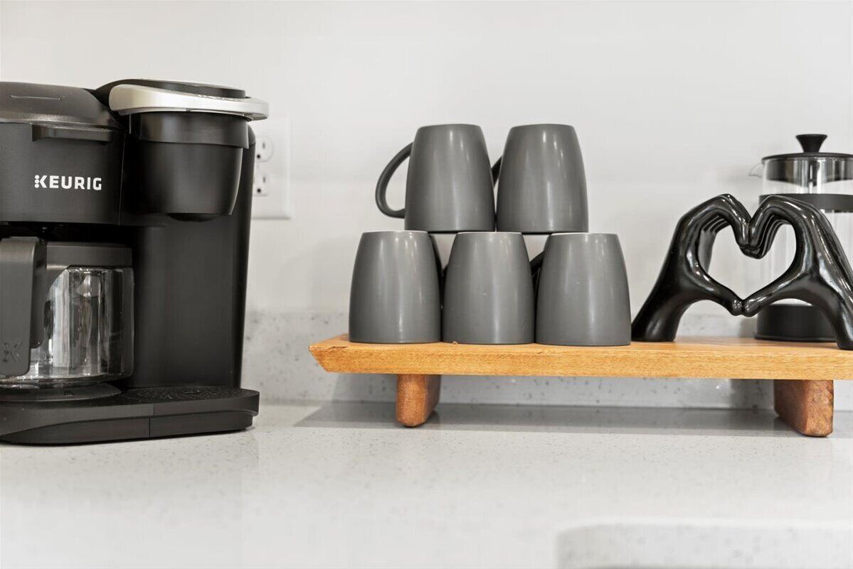 Une machine à café Keurig noire est posée sur un comptoir de cuisine. Une étagère en bois, située au-dessus, supporte six tasses grises et un objet noir en forme de cœur.