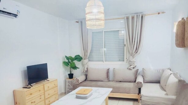 Ein Wohnzimmer mit einer Couch, einem Fernseher, einem Couchtisch, einer Pflanze und einem Fenster mit Vorhängen.