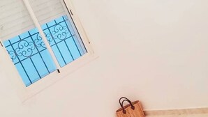 Ein Zimmer mit einem Bett, einem kleinen Tisch, einem Fenster mit Metallgitter und einer Wandleuchte.