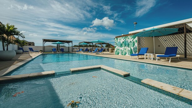 Ein Swimmingpool mit blauen Liegestühlen und Sonnenschirmen, umgeben von einer Holzterrasse und Palmen.