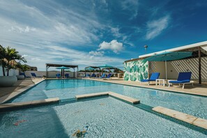 A swimming pool with blue lounge chairs and umbrellas, surrounded by a wooden deck and palm trees. - LUX Villas by Bocobay (Noord)