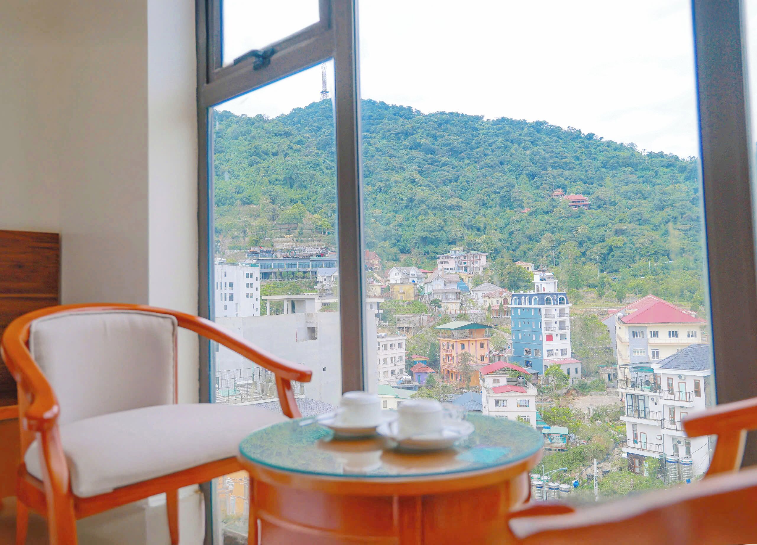 A room with a large window offering a view of a hillside with buildings. There is a round table with a teapot and cups, and two wooden chairs.