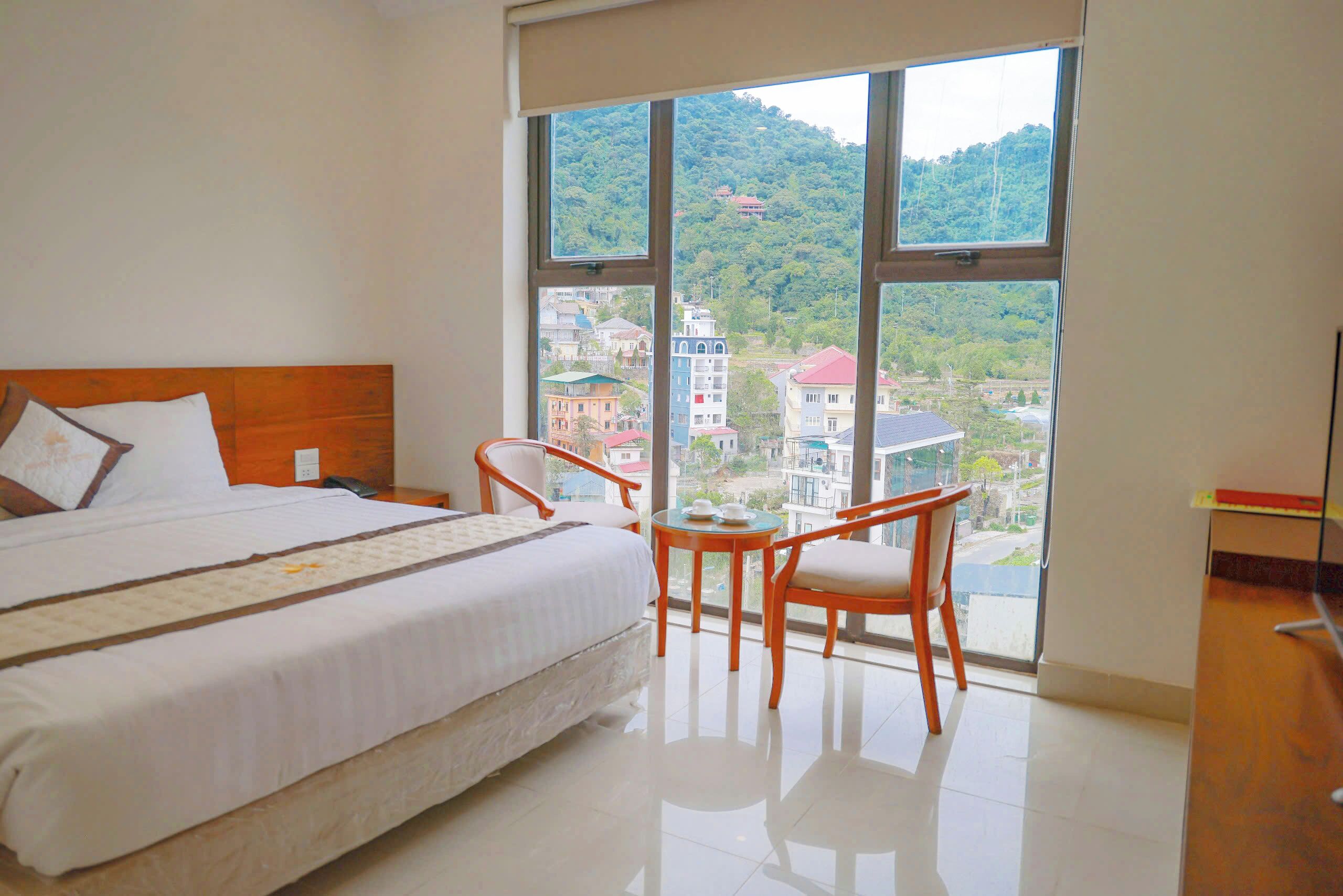 A hotel room with a large bed, two chairs, a small table, and a view of a cityscape through large windows.