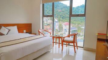 A hotel room with a large bed, two chairs, a small table, and a view of a cityscape through large windows.