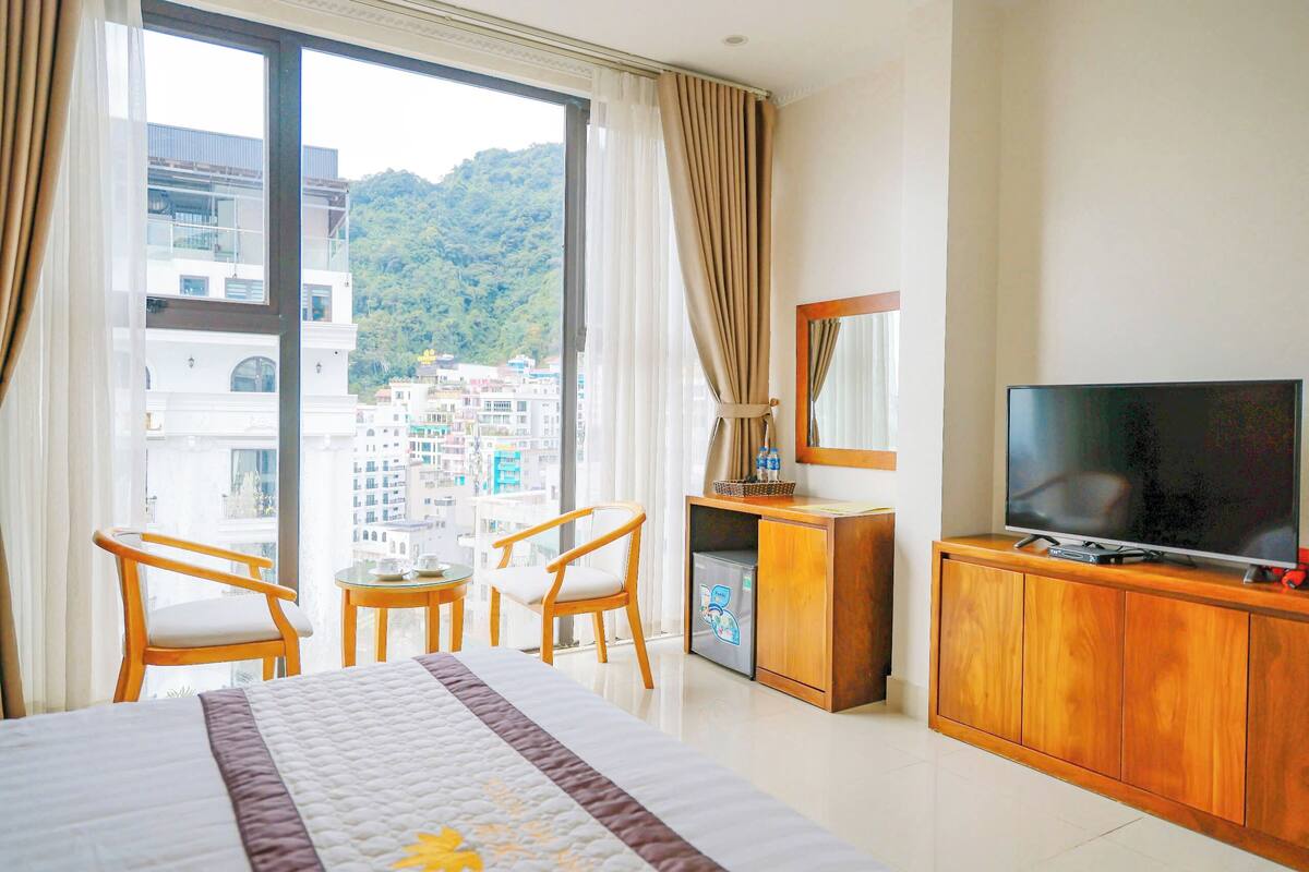 Habitación de hotel con una cama grande, un televisor en un mueble de madera, una pequeña mesa con sillas y vistas a un paisaje urbano a través de ventanas amplias.