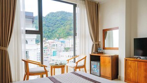 A hotel room with a large bed, two chairs, a small table, a television, and a view of a cityscape through a large window. - Ngan Ha Hotel Panorama View (Tam Dao)