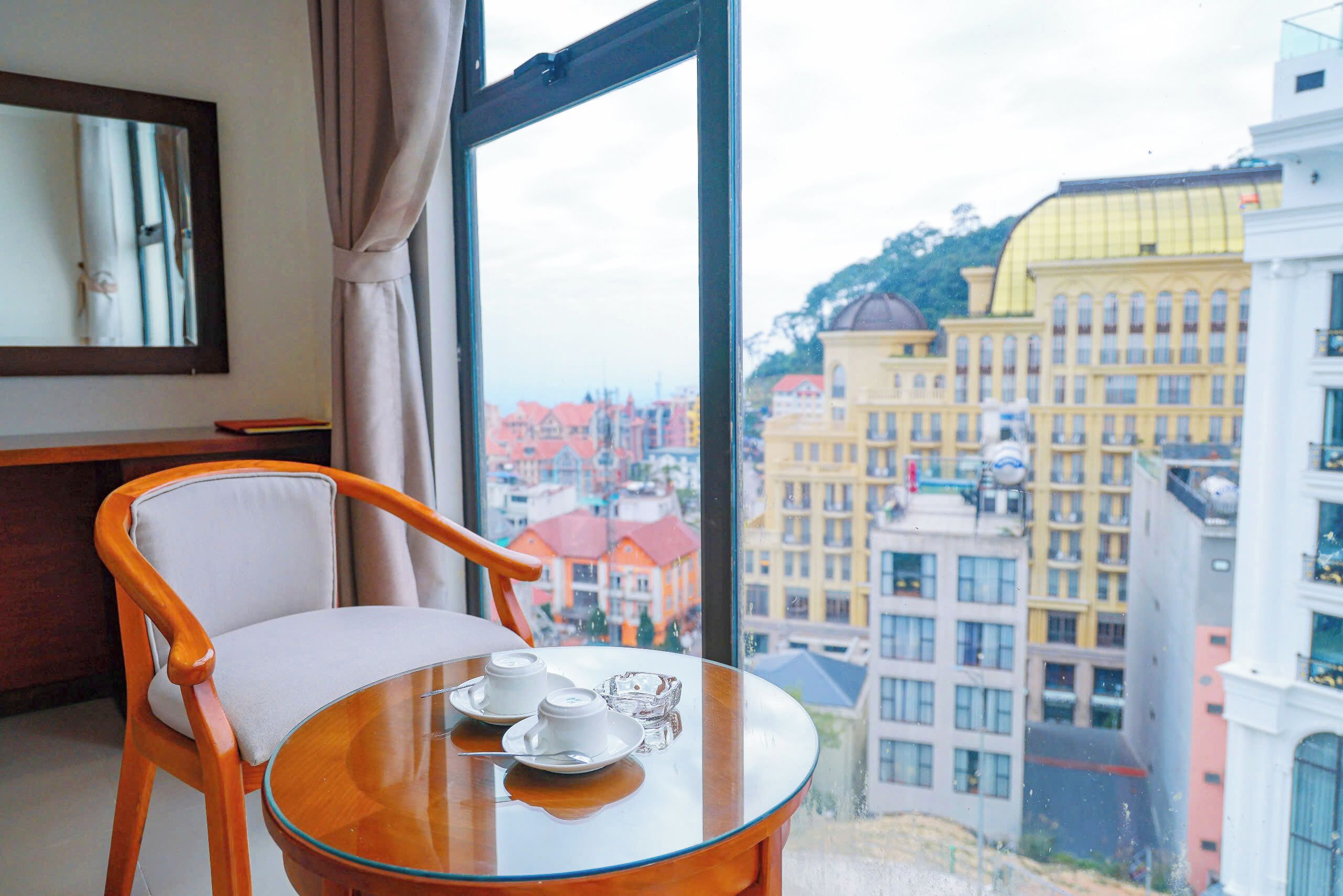 A hotel room with a round glass table, a wooden chair, and a view of city buildings.