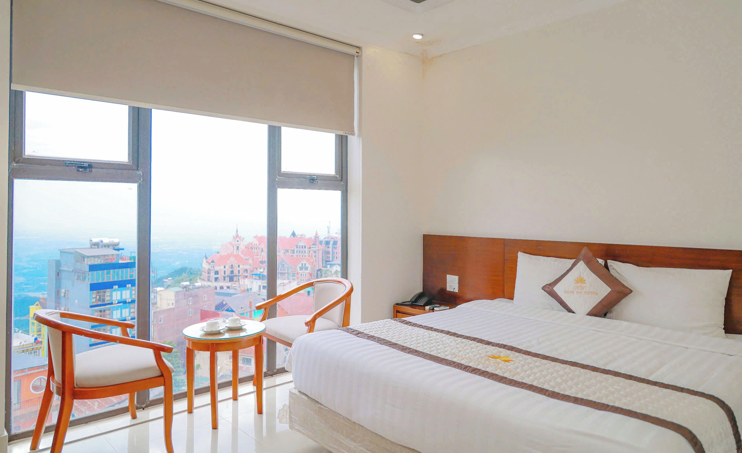 A hotel room with a large bed, a wooden chair, a small table with a teapot, and a city view through large windows.