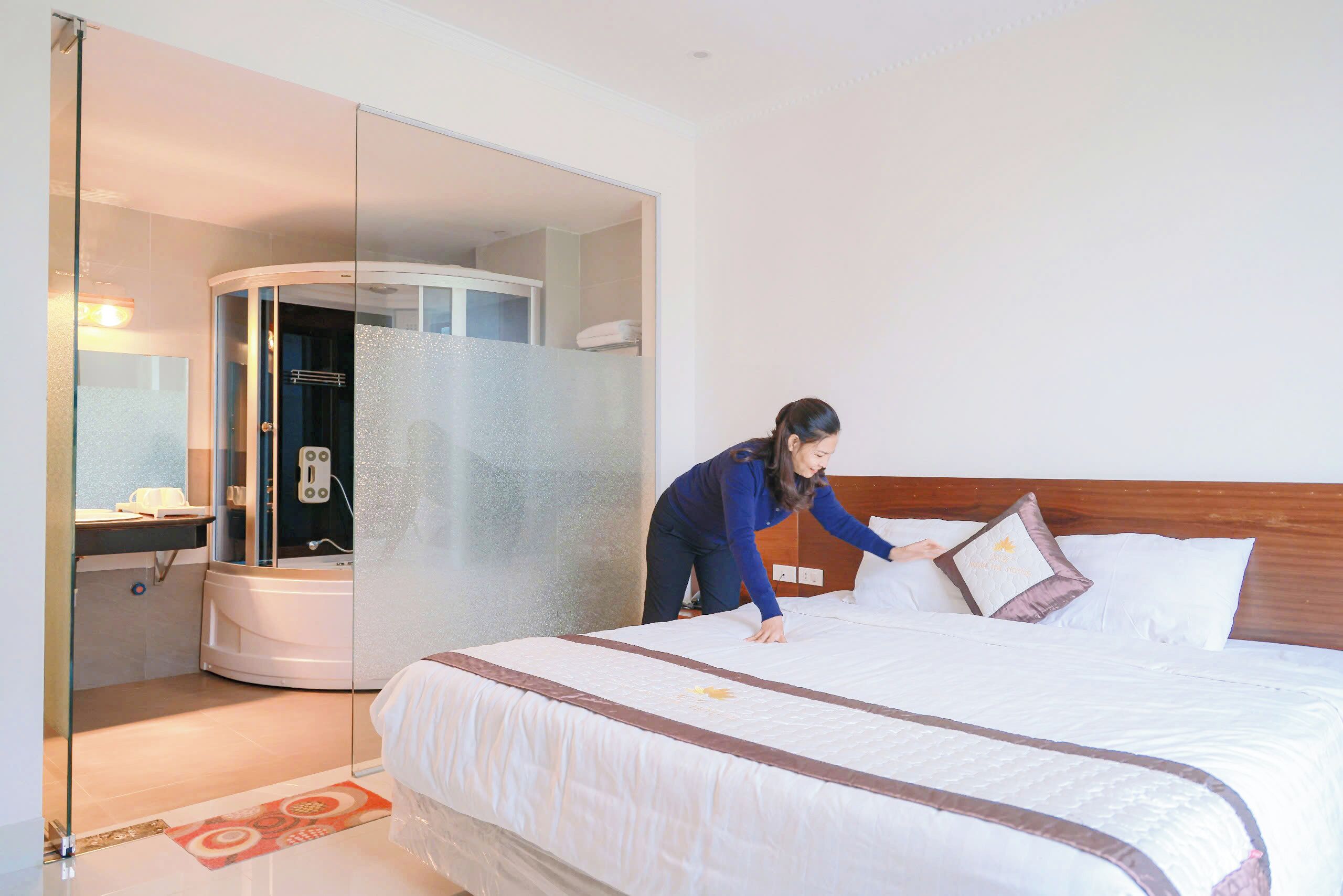 A hotel room with a bed, a glass partition, a bathroom with a shower, and a small desk with a chair.