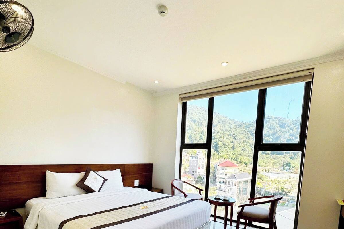 Habitación de hotel con una cama grande, una mesita de noche, una silla y vistas a un paisaje urbano a través de amplios ventanales.