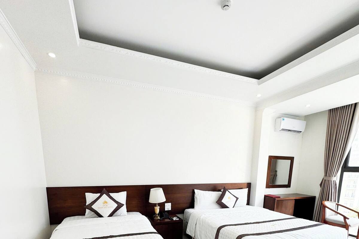 Habitación de hotel con dos camas, una mesita de noche, una lámpara, un equipo de aire acondicionado y una ventana con cortinas.