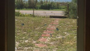 A view through a window of a paved pathway leading to a road, with a grassy area and a body of water in the background. - Stay in a Historic Beachfront Gem with Unmatched Ocean Views (Port St. Joe)