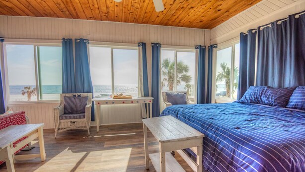 A room with a wooden ceiling, large windows with curtains, a bed with a blue bedspread, a small table, and a chair. - Stay in a Historic Beachfront Gem with Unmatched Ocean Views (Port St. Joe)