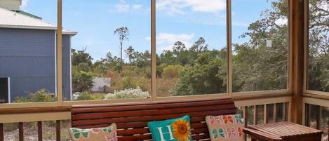 Un banco de madera con cojines florales y vistas a una casa azul y abundante vegetación a través de amplios ventanales.