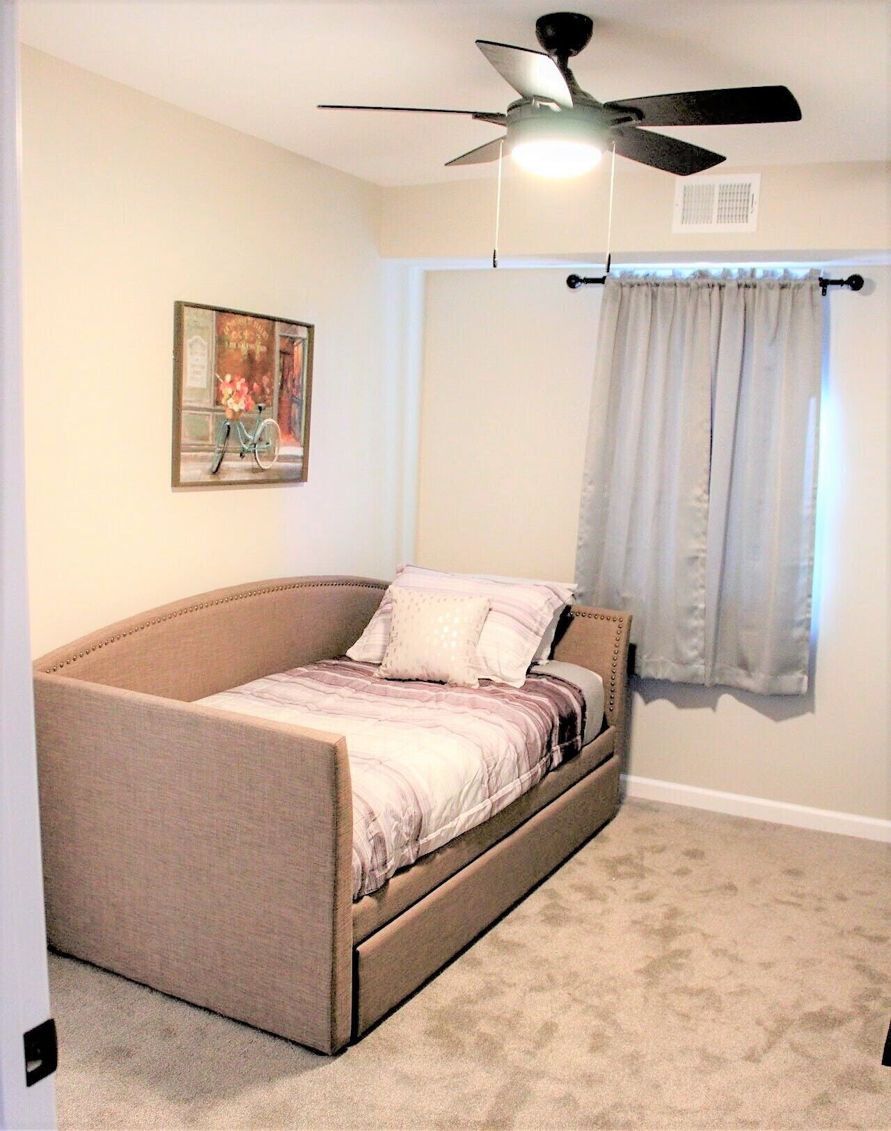 A bedroom with a single bed, a ceiling fan, a window with curtains, and a painting on the wall.