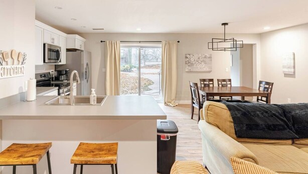 A modern kitchen with a central island, a dining area with a wooden table and chairs, and a cozy seating area with a sofa and cushions. - Guesthouse 3BDR Suite (Overland Park)