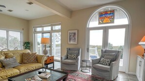 Un salon comprenant un canapé, deux fauteuils, une table basse et un tapis. Il y a une fenêtre ornée de vitraux et une porte donnant sur l’extérieur.