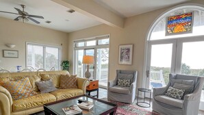 Un salon avec un canapé jaune, une table basse en verre et deux fauteuils. On y trouve une grande fenêtre avec vue, un ventilateur de plafond et un tapis.