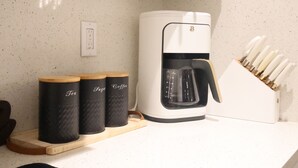 Una cucina con una macchina da caffè, due contenitori etichettati 'Tè' e 'Zucchero e Caffè', e un portachiodi.