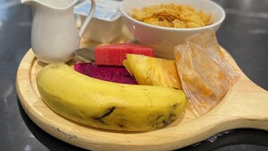 Een houten dienblad met een banaan, pitahaya, watermeloen en een kom ontbijtgranen op een tafel.