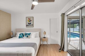A modern bedroom with a bed, bedside lamps, a ceiling fan, and a view of a swimming pool through glass doors.
