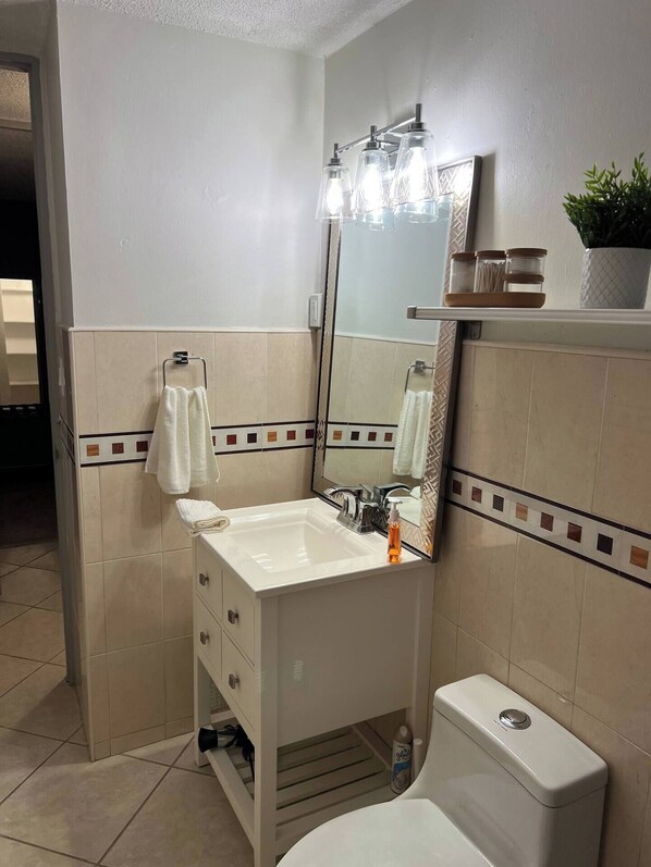 A bathroom with a white sink, mirror, and toilet. There is a towel rack with a towel hanging on it, and a shelf with a plant and toiletry jars.