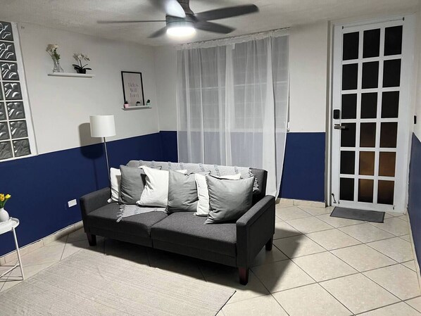 Una habitación con un sofá de gris oscuro, una puerta blanca, un ventilador de techo y una ventana con cortinas traslúcidas.