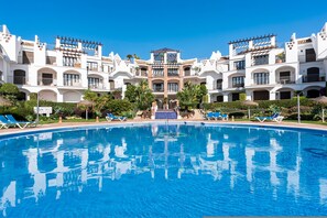 A large swimming pool with blue water, surrounded by white buildings with balconies and a central staircase leading to a covered area. - Los Arqueros Sunny Balcony With Pool - Ref M52 (Benahavis)