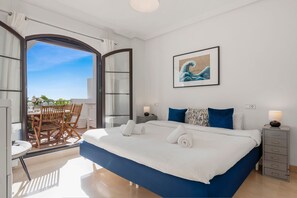 A modern bedroom with a large bed, a view through an open door to a balcony, a framed artwork on the wall, and a bedside table with a lamp. - Los Arqueros Sunny Balcony With Pool - Ref M52 (Benahavis)
