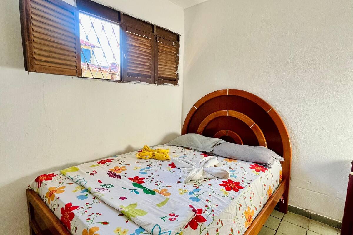 Una cama individual con colcha floral, cabecero de madera y ventana con contraventanas de madera.