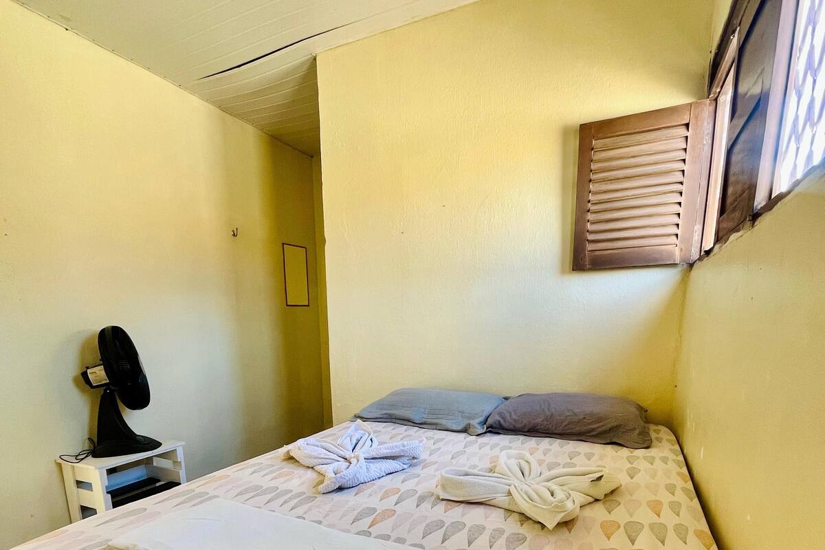 Una cama bien hecha con colcha estampada y toallas, una mesita de noche con lámpara, y una ventana con persianas.