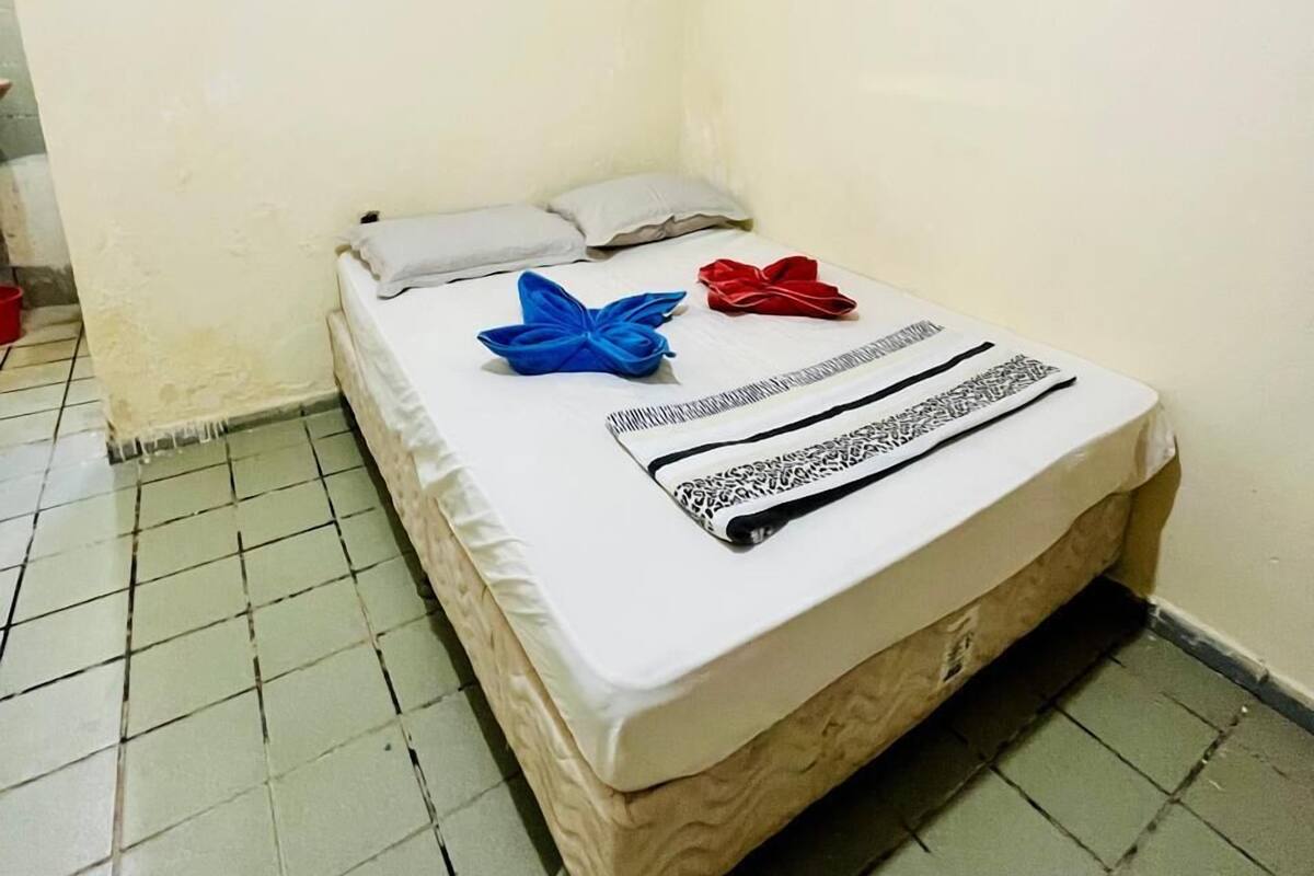 Una habitación pequeña con cama individual, pared blanca sencilla y suelo de baldosas.