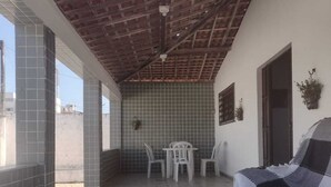 A covered patio area with tiled flooring, a white table, two white chairs, a sofa, and a potted plant. - Harmô Hostel Cultural (Conde)
