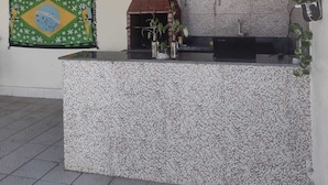 A covered outdoor area with a brick fireplace, a tiled floor, a wall-mounted shelf with plants, and a Brazilian flag. - Harmô Hostel Cultural (Conde)