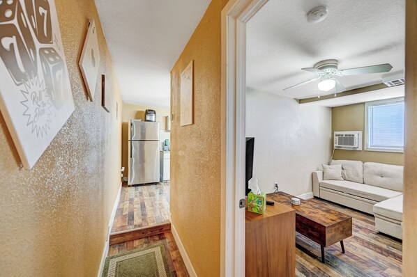 A hallway leading to a living room with a ceiling fan, a refrigerator, and a wooden coffee table.
