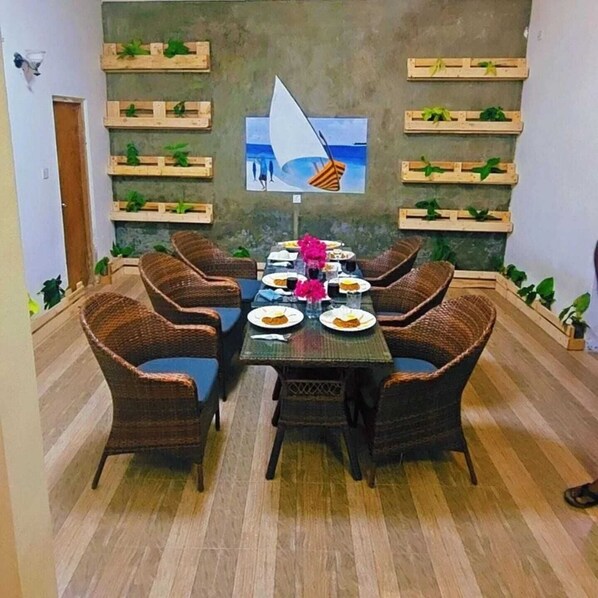 A dining area with a long table, wicker chairs, and wooden shelves with plants. - Dhontakuru Guesthouse (Dhidhdhoo)