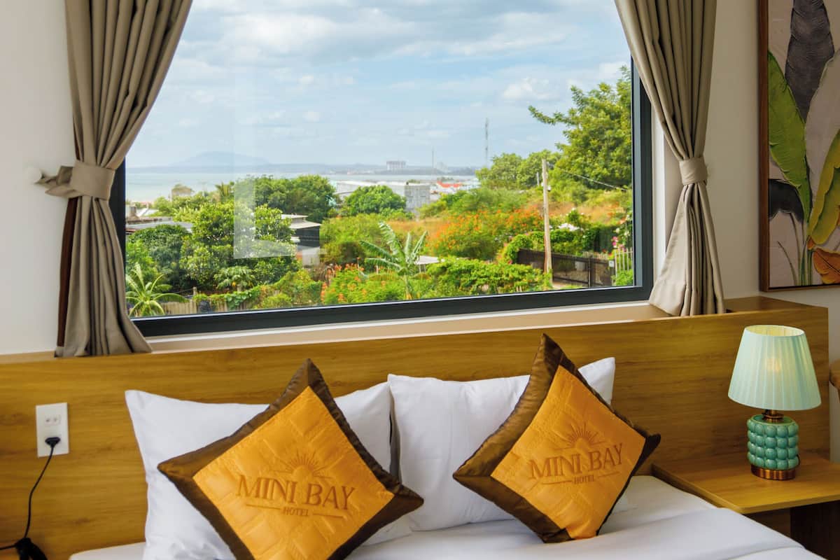 A hotel room with a bed, two pillows with 'MINI BAY HOTEL' text, a nightstand with a lamp, and a window with curtains.