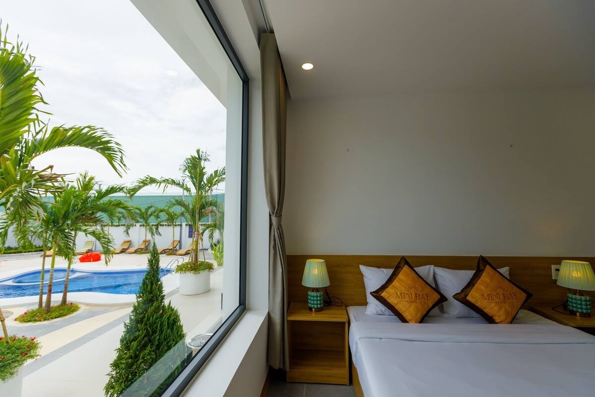A hotel room with a bed, nightstands, and a view of a swimming pool and palm trees.