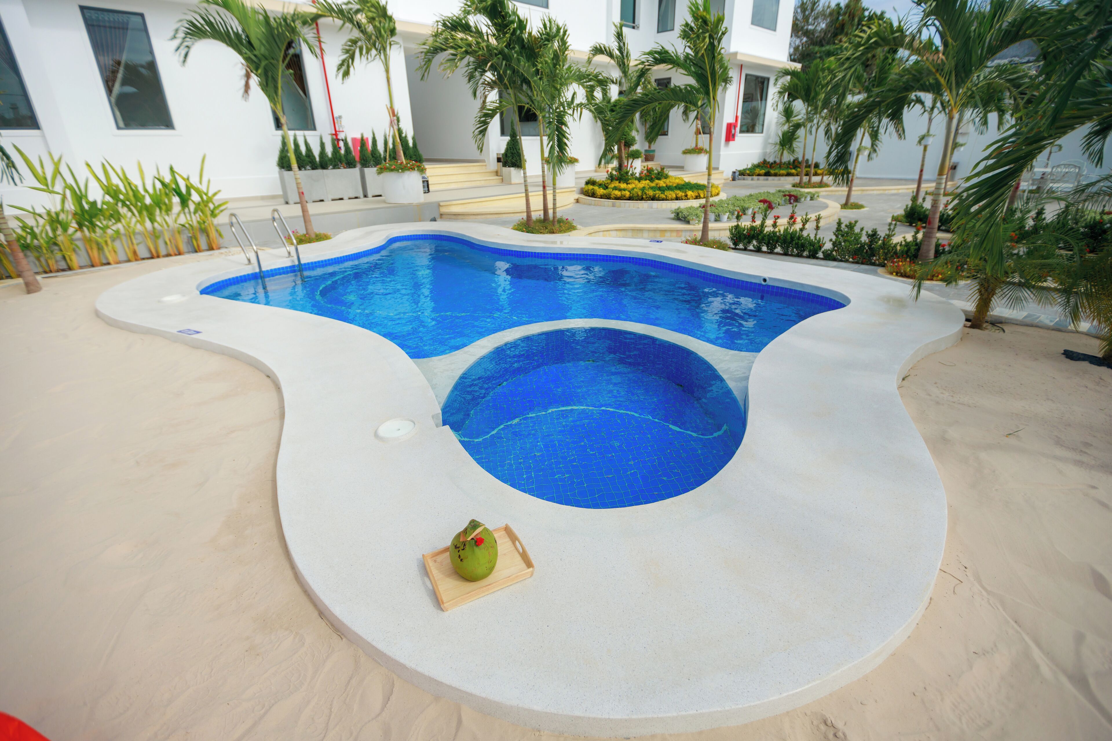 Une piscine à la forme originale, entourée d’une zone sablonneuse et agrémentée de différentes plantes.