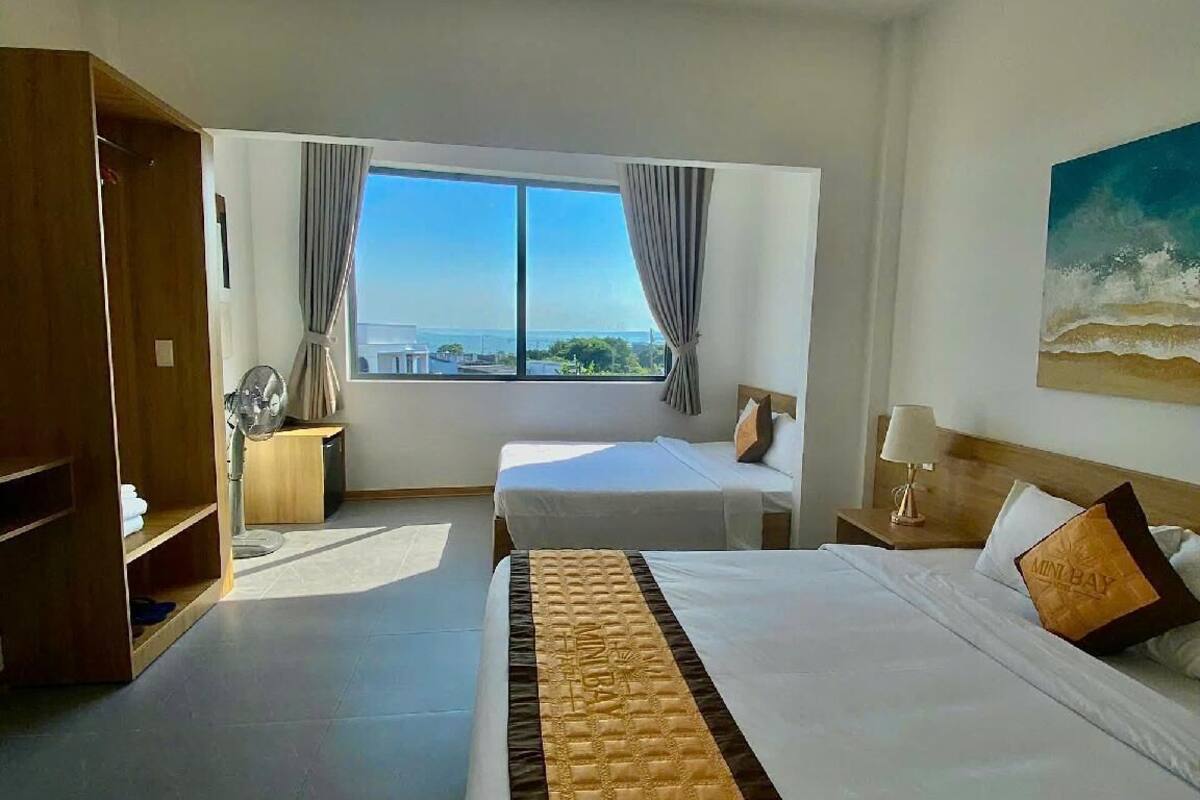 A hotel room with two beds, a large window with a city view, a wooden wardrobe, and a painting of a beach scene.